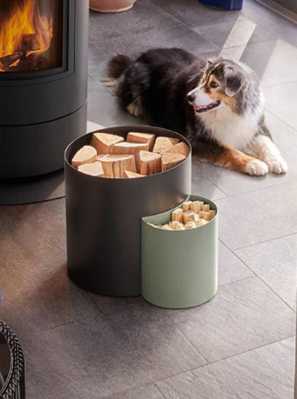 Range-bûches DIXNEUF gris et vert sauge près d’un poêle à bois allumé, avec un chien allongé à côté.